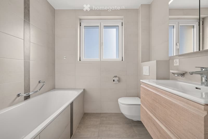 Modern bathroom with a bathtub, toilet, sink, and large mirror. Light-colored tiles and wooden vanity.