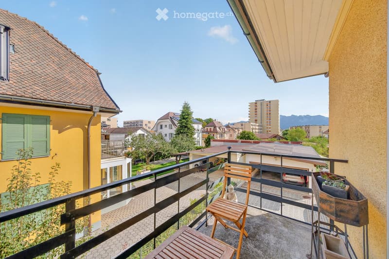 Balcony with wooden chair, table, and potted plant, overlooking a residential area with houses and mountains in the background.