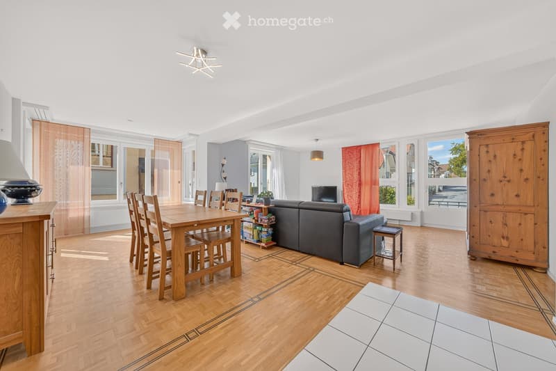 Bright living and dining area with wooden flooring, large windows, a grey sofa, a wooden dining table with chairs, a television, side table, and a cabinet.
