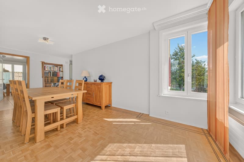 Bright dining room with wooden flooring, large windows, a wooden dining table surrounded by chairs, a wooden cabinet with a lamp and decorative items, and a bookshelf filled with books.