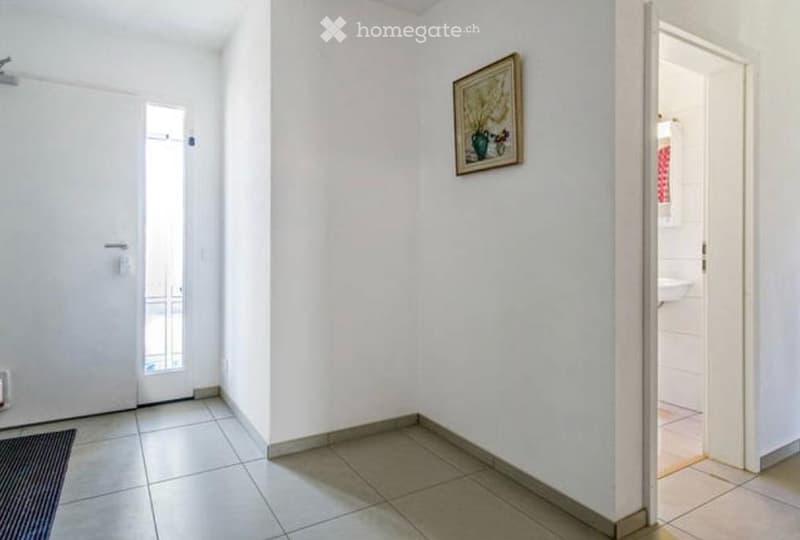 Empty hallway with white walls, tiled floor, and a small rug near the entrance door. A framed picture hangs on one wall, and an open doorway leads to another room.
