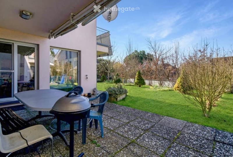 Outdoor patio with a table, chairs, and a grill, overlooking a well-maintained lawn and garden area.