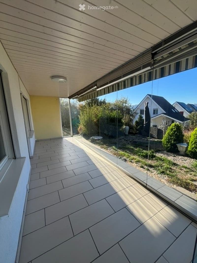 Spacious balcony with tiled flooring, large glass railing, and a view of the garden and neighboring houses.
