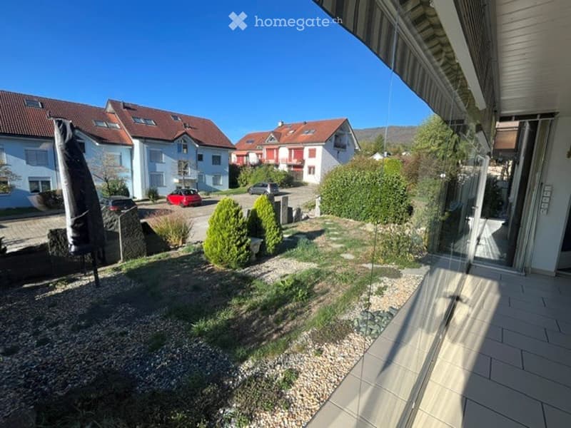 Well-maintained garden area with gravel pathways, small shrubs, and a patio connected to a modern house. The surrounding neighborhood features similar houses with red-tiled roofs and white facades.