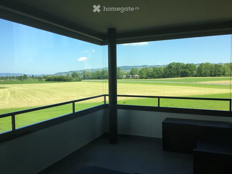 Balcony with a view of a grassy field and distant hills, featuring a metal railing and a small bench.