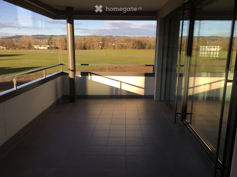 Modern balcony with tiled floor, glass railings, and a panoramic view of a green field and distant landscape.