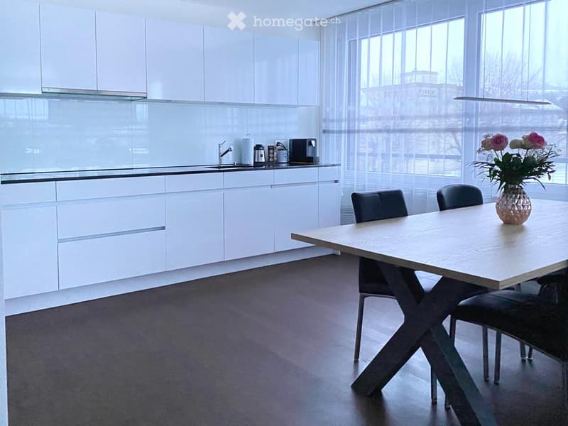 Modern kitchen with white cabinets, a sink, a coffee maker, and a dining area with a table and chairs. A vase with flowers is placed on the table.