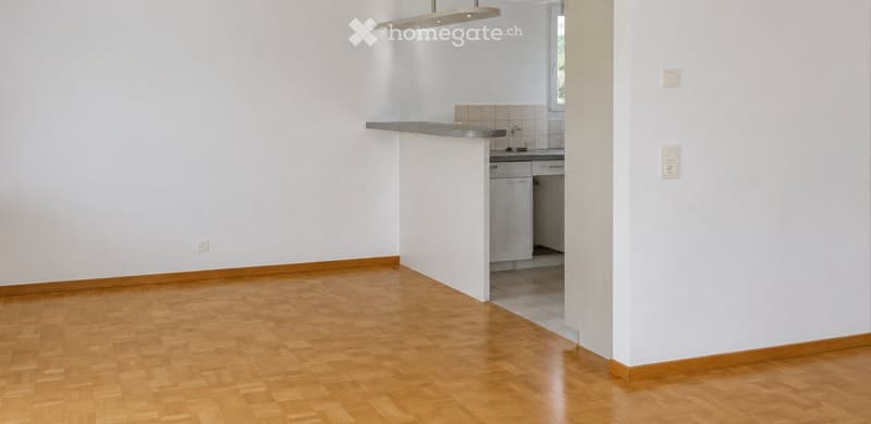 Empty room with wooden flooring, white walls, and a small kitchen area with cabinets and a countertop.