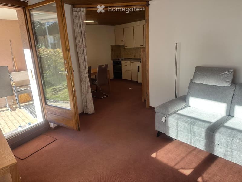Bright living room with a grey sofa, dining area with table and chairs, and an adjacent kitchen with white cabinets and an oven. Large sliding glass door leads to a patio.