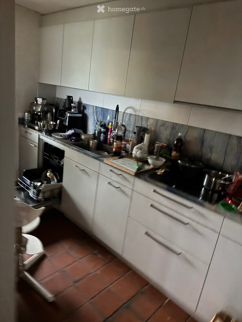 Modern kitchen with white cabinets, stainless steel appliances, and a tiled floor. The counter is cluttered with various kitchen items including a coffee maker, cutting board, frying pan, and other utensils.