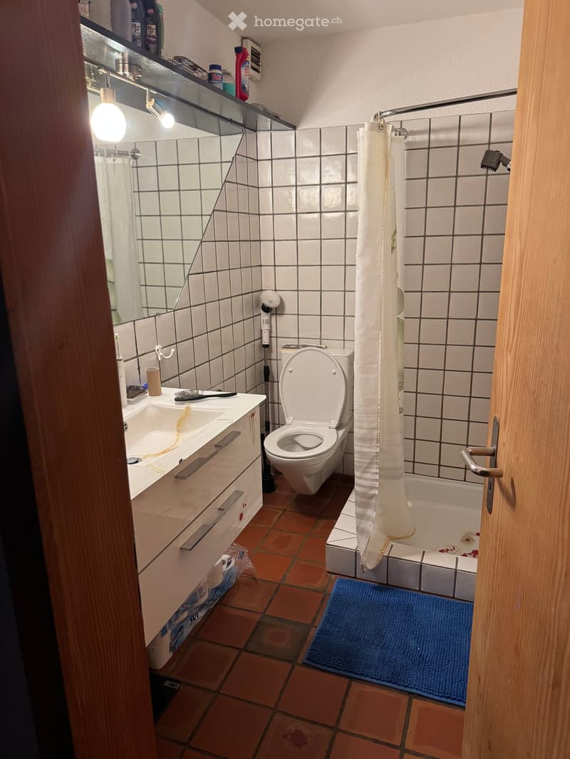 Small bathroom with white tiled walls, a toilet, a sink with a mirror, a bathtub with a shower curtain, and a blue bath mat on the floor.