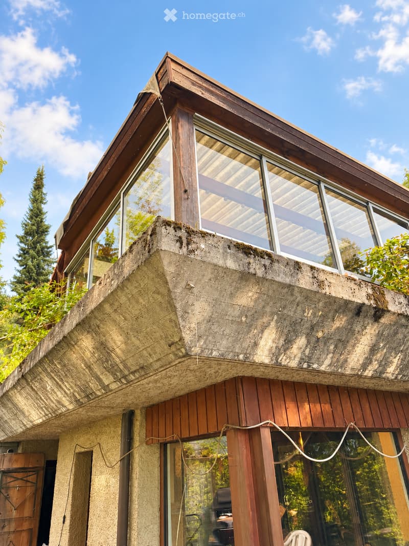 Modern house with a unique, angular design featuring a combination of concrete and wooden elements. The roof has a distinctive, sloped structure with large windows, and the facade includes a mix of textured concrete and wooden panels.