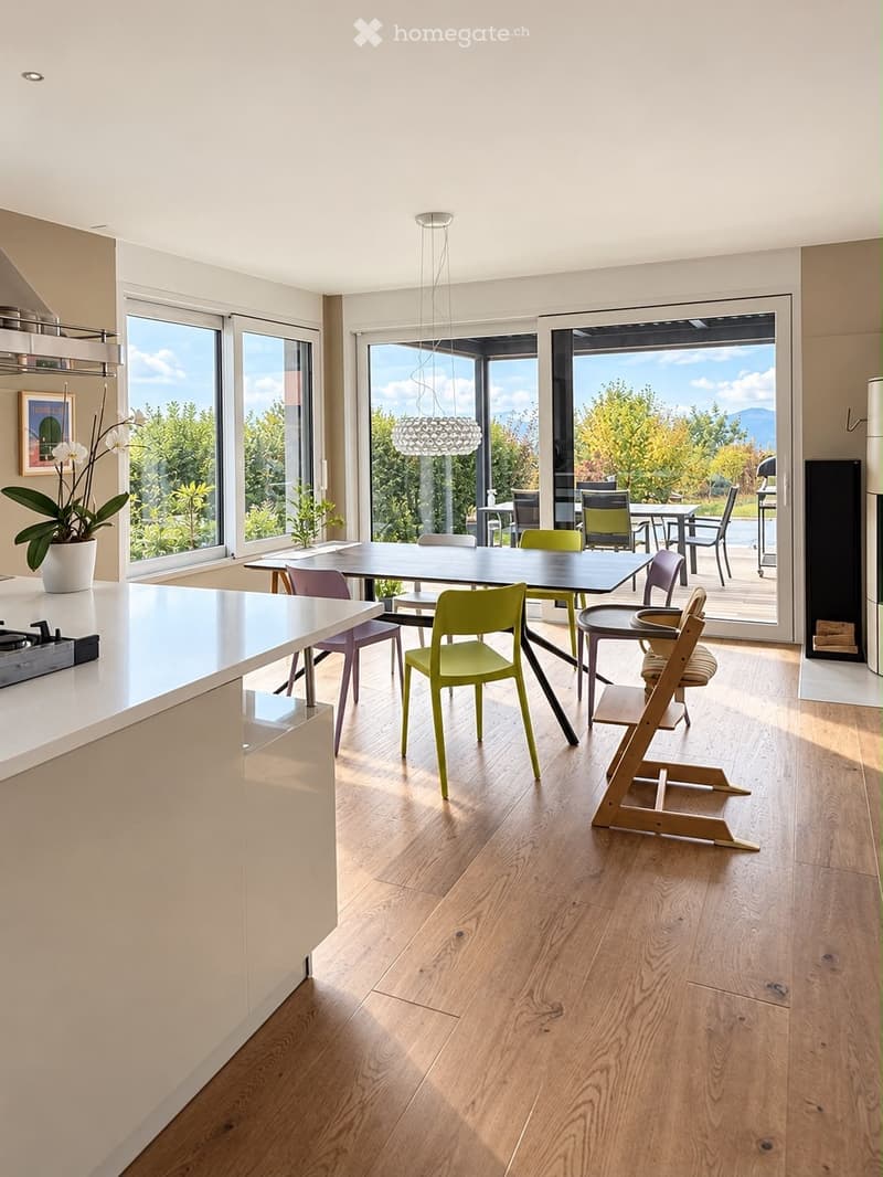 Bright, modern kitchen and dining area with light wood flooring, white cabinetry, and large windows overlooking a scenic outdoor space. Features a central dining table with colorful chairs, a high chair, and a kitchen island with a gas stove.