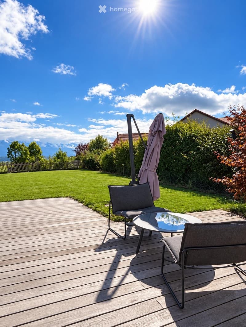 Wooden deck with two grey chairs and a round glass table, umbrella folded nearby, surrounded by a well-maintained lawn and greenery.