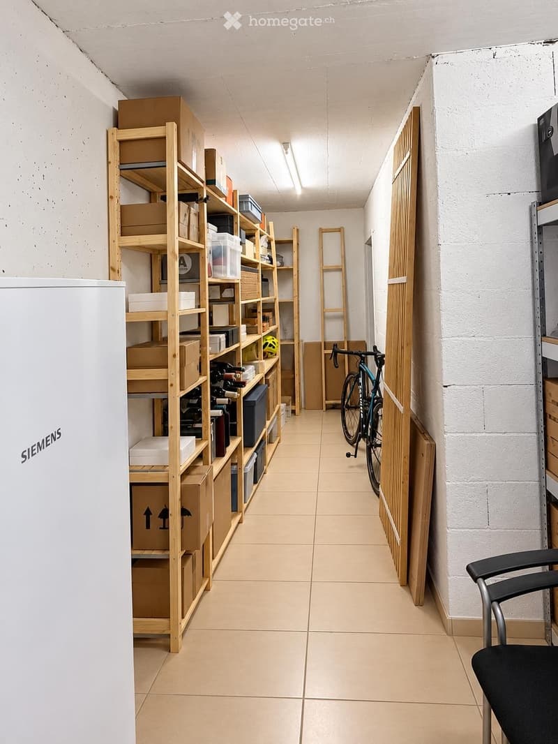 A narrow storage room with wooden shelves filled with boxes and items, a bicycle leaning against the wall, and a black chair in the foreground.