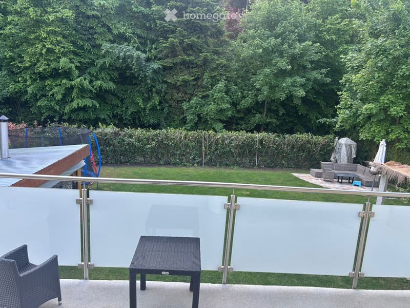 Balcony with a modern glass railing, featuring a wicker chair and a small table, overlooking a well-maintained garden with seating area and umbrella.