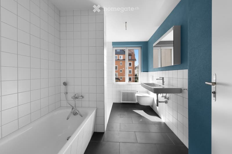 Modern bathroom with white tiled walls, a bathtub, toilet, sink, mirror, and a window offering a view of the street.
