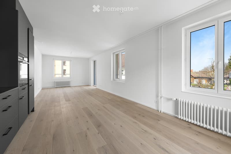 Empty room with light wooden flooring, white walls, multiple windows, and radiators. No furniture or furnishings present.