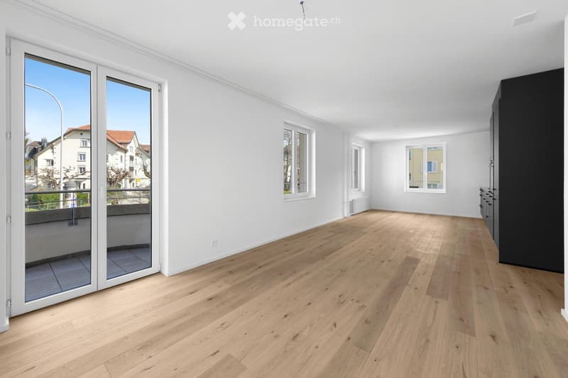 Empty room with light wooden flooring, white walls, large windows, and a sliding glass door leading to a balcony.