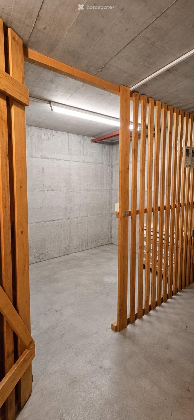 Concrete-walled corridor with wooden slatted partitions and overhead lighting, leading to a garage area.