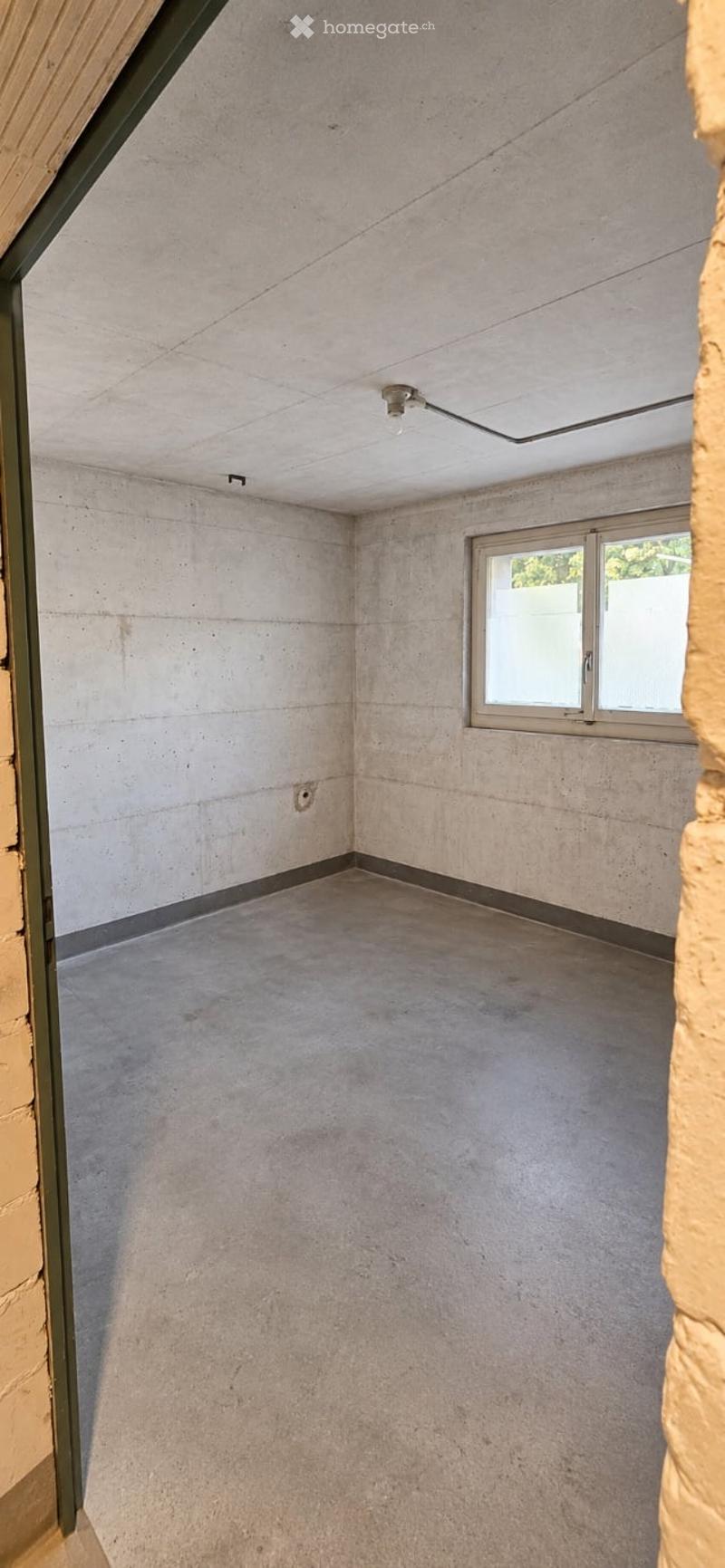 Empty room with concrete walls and floor, a single window with metal frame, and a ceiling light fixture.
