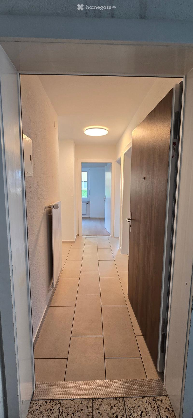 Clean, well-lit hallway with tiled flooring, white walls, and a recessed ceiling light. Features a wooden door on the right and a radiator on the left, leading to another room with visible furniture.