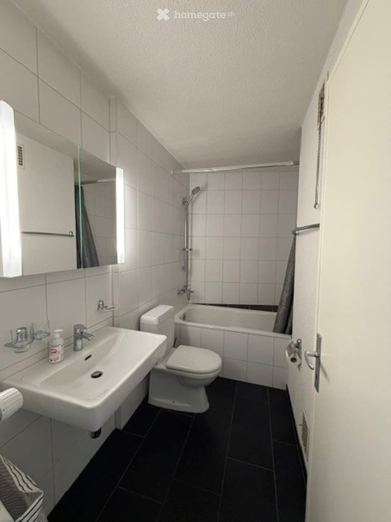 Modern bathroom with white tiles, black floor, sink, toilet, bathtub, and shower. Features a mirror and cabinet above the sink.