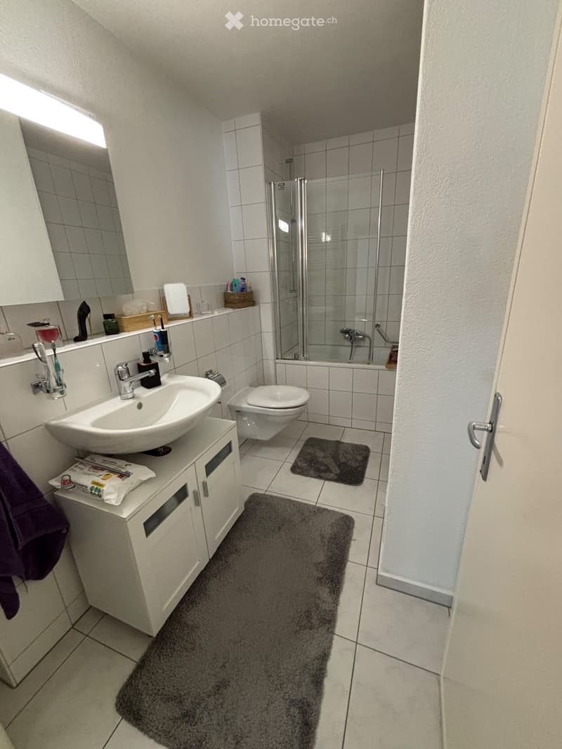 Modern bathroom with white tiles, a sink with a modern faucet, a toilet, a shower stall with glass doors, a mirror, a cabinet, and a towel on the floor.