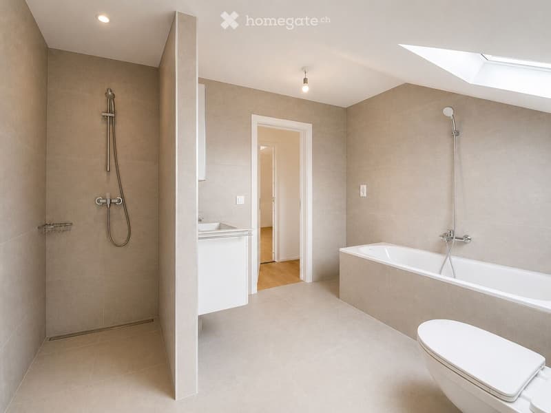 Modern bathroom with beige tiles, a bathtub, toilet, sink, and showerhead. Features include a skylight, recessed lighting, and a small shelf.