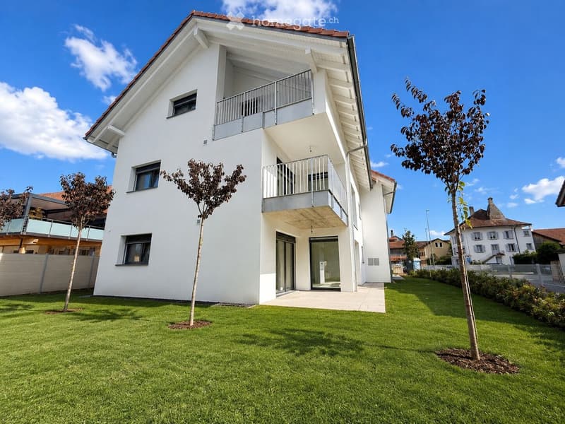 Modern two-story house with white exterior, large windows, and balconies. The house features a well-maintained lawn with small trees and is situated in a residential area with neighboring houses visible in the background.