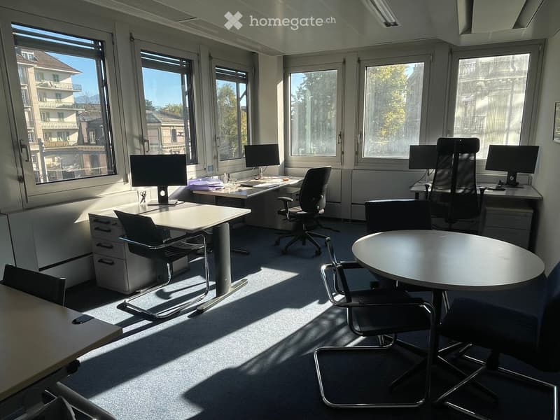 Bright office space with large windows, desks, chairs, computer monitors, and a round meeting table.