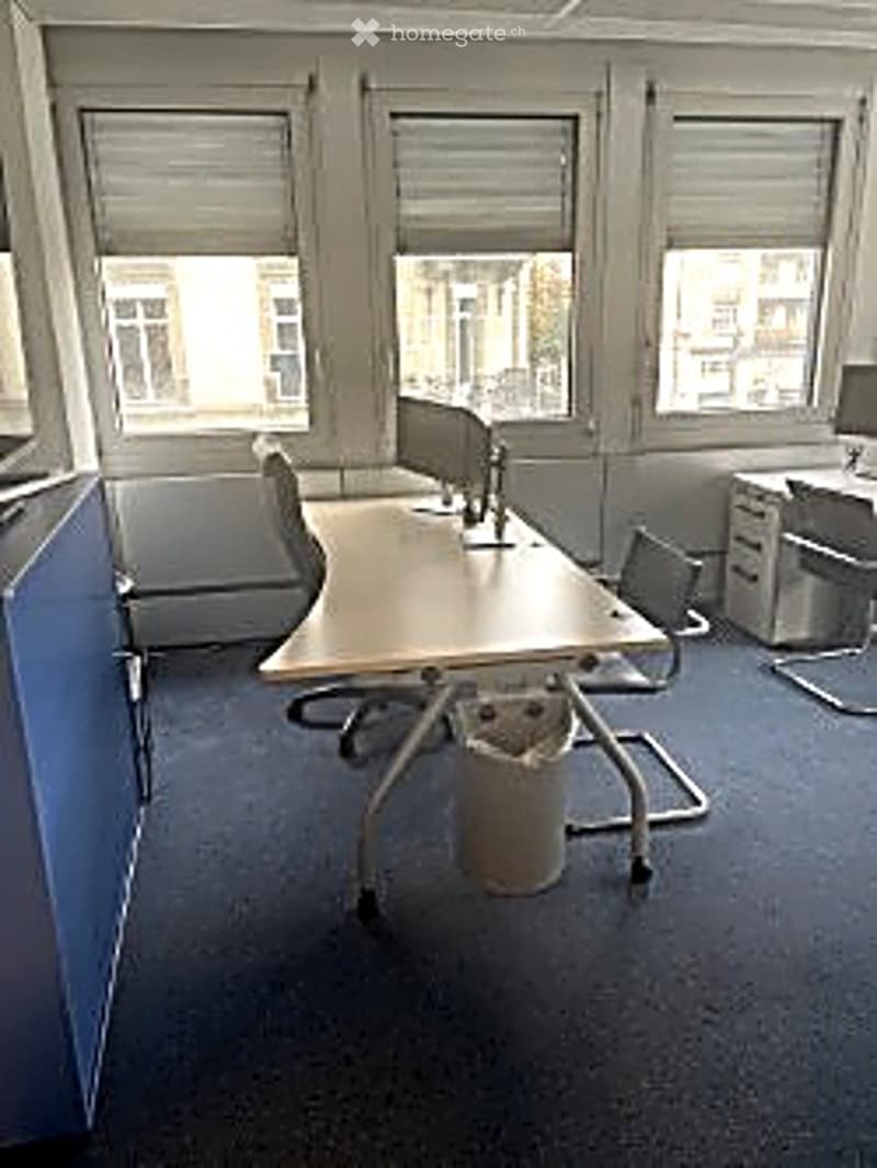 Empty office space with large windows, desks, chairs, and a filing cabinet.
