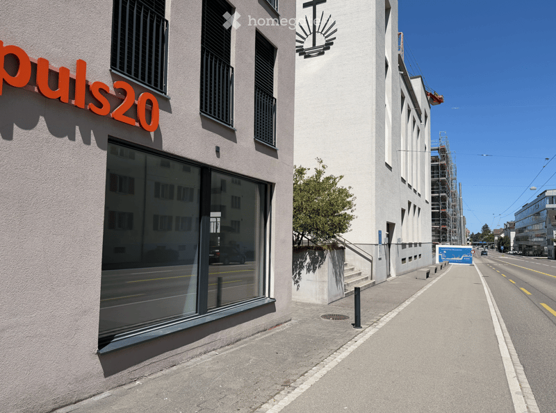Modern building facade with large windows and a sign reading "puls20" in orange letters. The building has a clean, contemporary design with a small staircase leading up to the entrance.