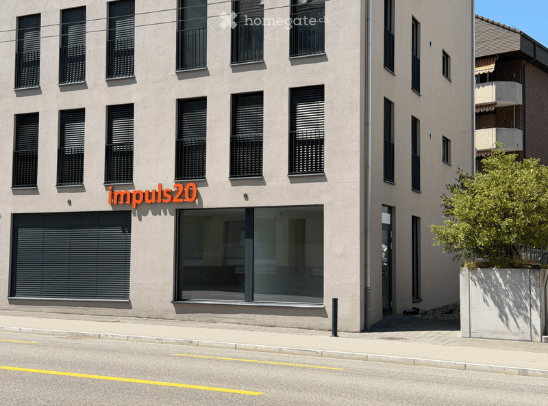 Modern multi-story building with large windows, black shutters, and a glass entrance. The facade is light-colored with a clean, contemporary design.
