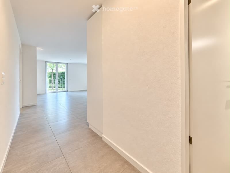 Empty hallway with tiled flooring, white walls, and a sliding glass door leading to a balcony area with outdoor views.