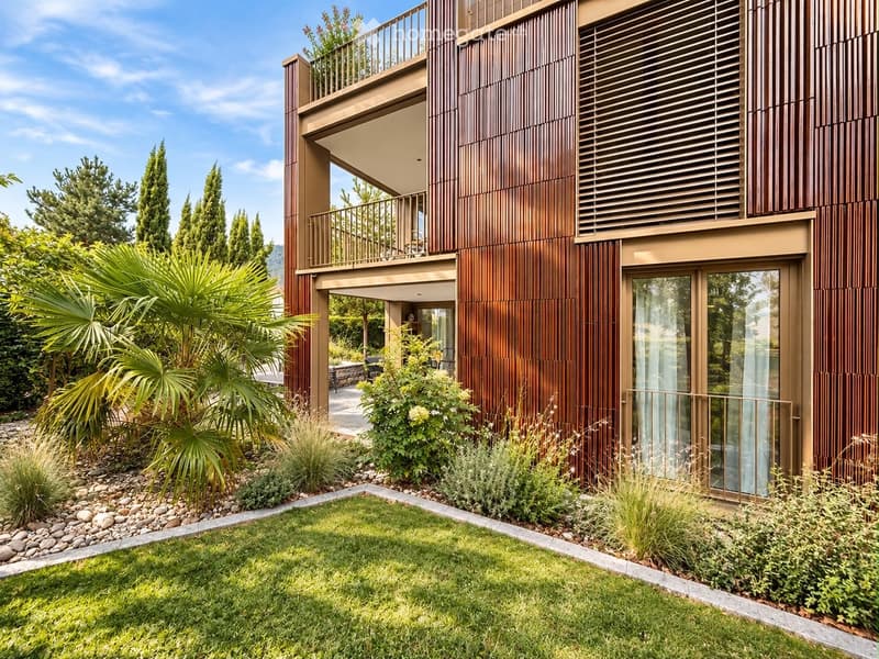 Modern two-story building with wooden facade, large windows, and a balcony. The surrounding area features a well-maintained garden with palm trees, shrubs, and a neatly trimmed lawn.