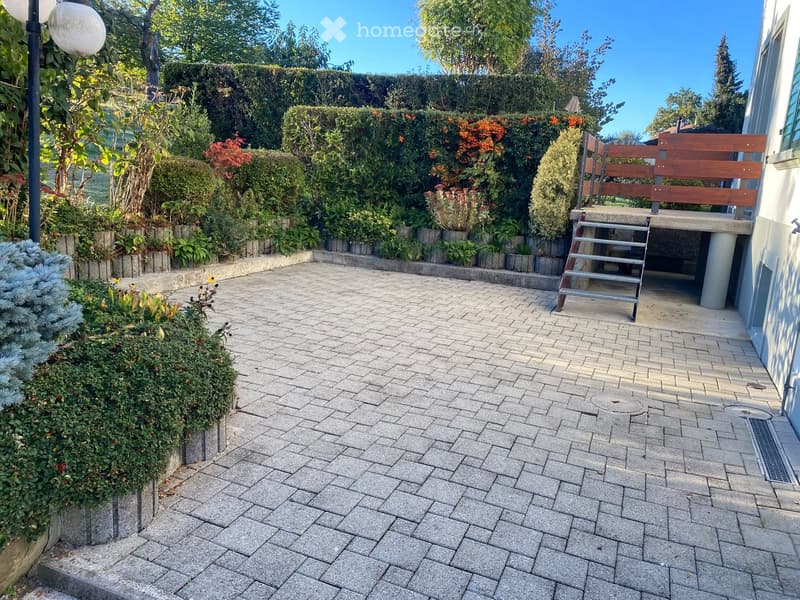 Well-maintained garden with a paved brick pathway, neatly trimmed hedges, flowering plants, and a wooden bench near the house.