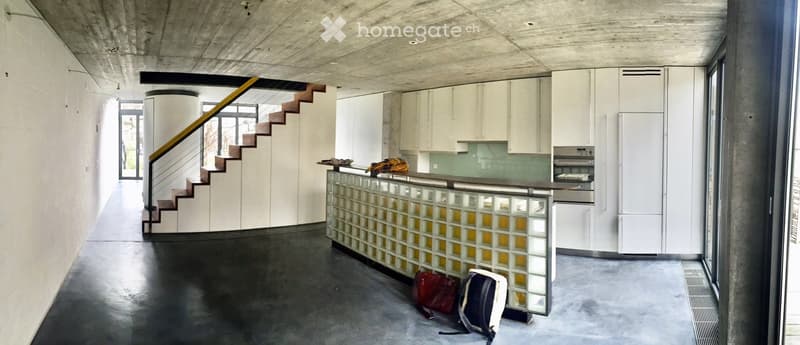 Modern, minimalist kitchen area with white cabinets, stainless steel appliances, and a glass block partition. A staircase leads upwards, and personal items like a handbag and backpack are placed on the floor.