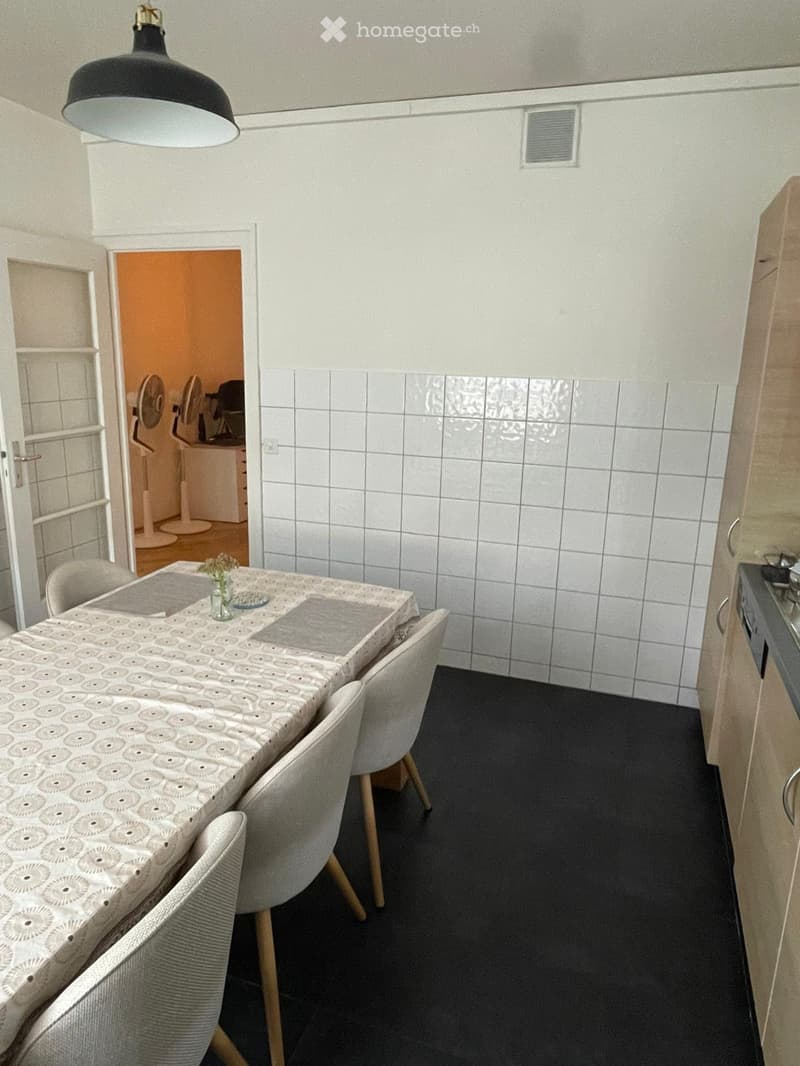 Dining room with a patterned tablecloth, four chairs, a fan in the adjacent room, and a small cabinet.