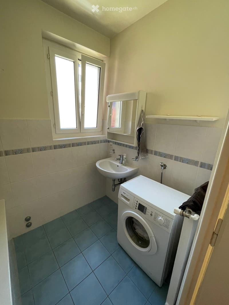 Bathroom with a sink, mirror, and a washing machine. Light-colored tiles and a window provide natural light.