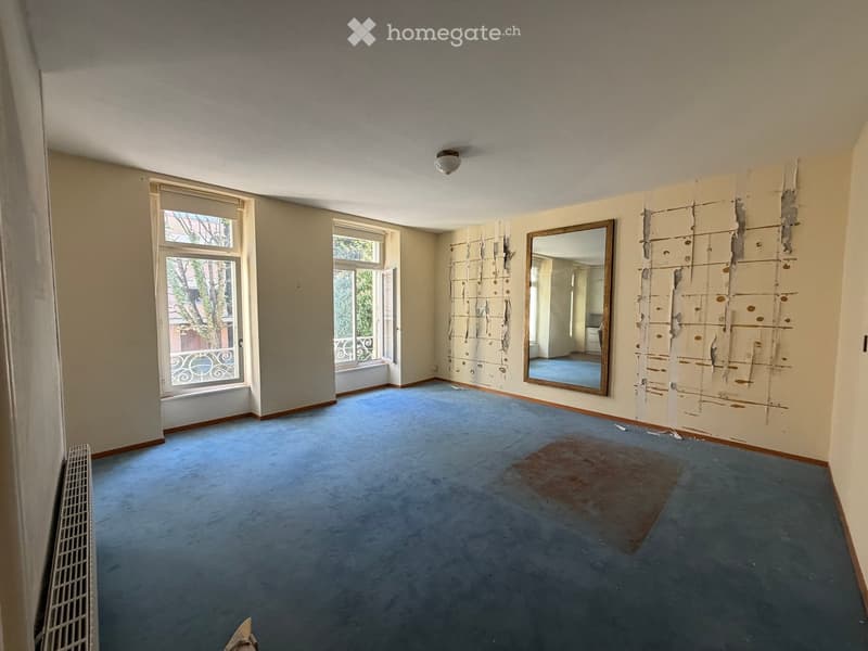 Empty room with blue carpet, exposed wall patches, large windows, and a mirror on one wall.