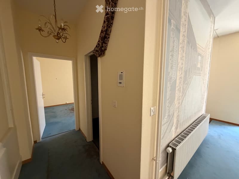 A narrow hallway with light-colored walls, blue carpeting, and a radiator on the right side. A chandelier hangs from the ceiling, and there are two open doors leading to other rooms.