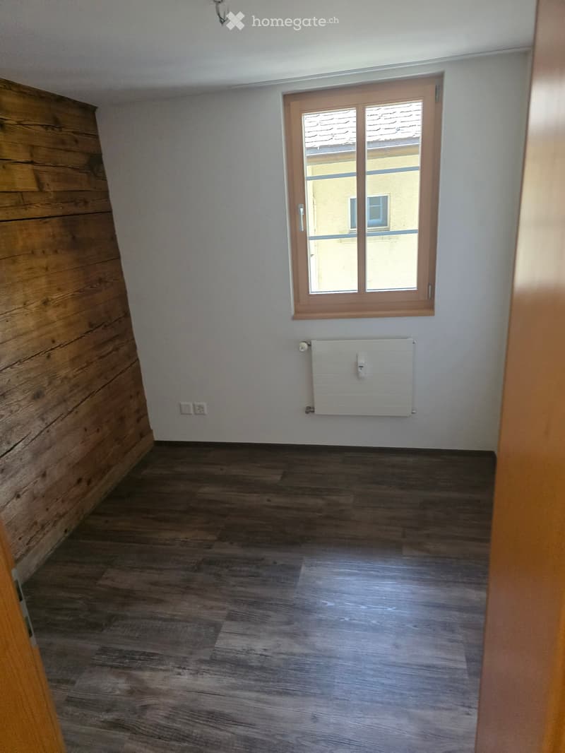 Empty room with wooden flooring, white walls, a window with wooden frame, and a radiator below it.