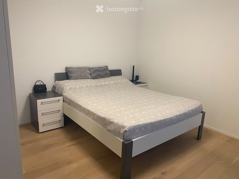 A modern bedroom with a single bed, nightstand, and handbag on the floor. The room features light-colored walls and wooden flooring.