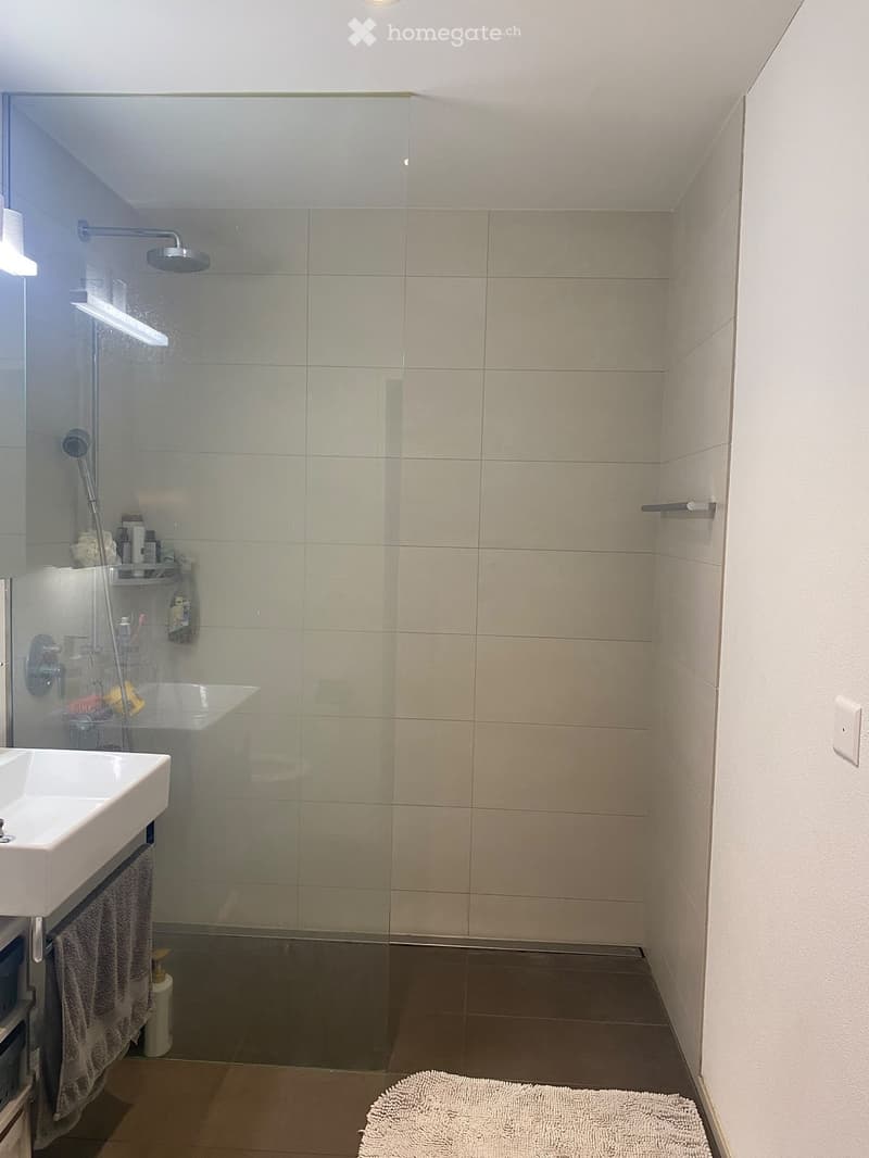 Modern bathroom with a white sink, glass-enclosed shower, beige tiles, and a towel on the floor.