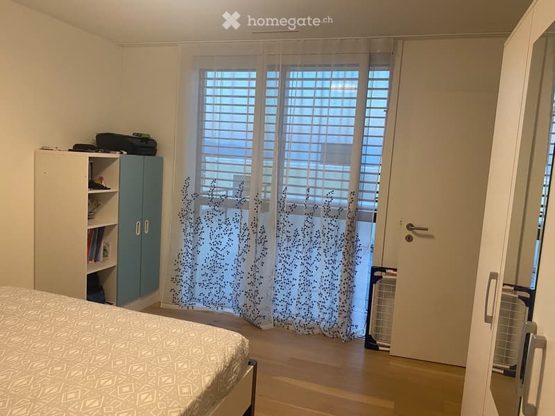 A tidy bedroom with a bed, a white cabinet with shelves, a printer on top, and sheer curtains with a leafy pattern covering the windows.