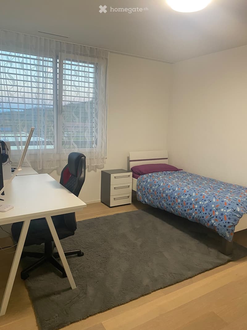 A small, well-lit bedroom with a single bed, desk, and chair. The bed has a colorful duvet and is positioned next to a window with sheer curtains. A desk with a laptop and a chair is placed near the window, and a small drawer unit is visible next to the b