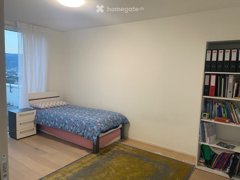 Cozy bedroom with a single bed, nightstand, bookshelf filled with books, and a scenic view through large windows.