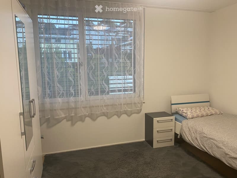 A tidy bedroom with a bed, nightstand, and a window with sheer curtains. The room features a grey carpet and white walls.