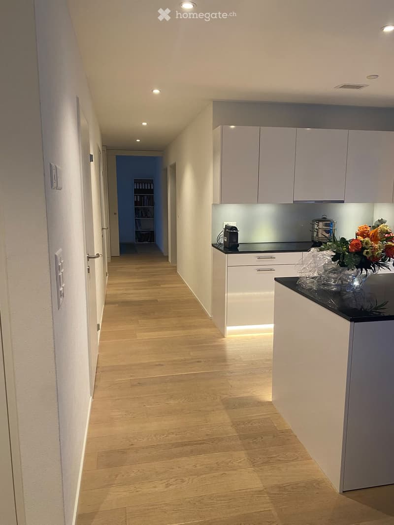 A modern hallway with light wooden flooring, white walls, and recessed lighting. A small kitchen area is visible on the right with white cabinets, a black countertop, and a vase with flowers. A bookshelf is visible in the background.